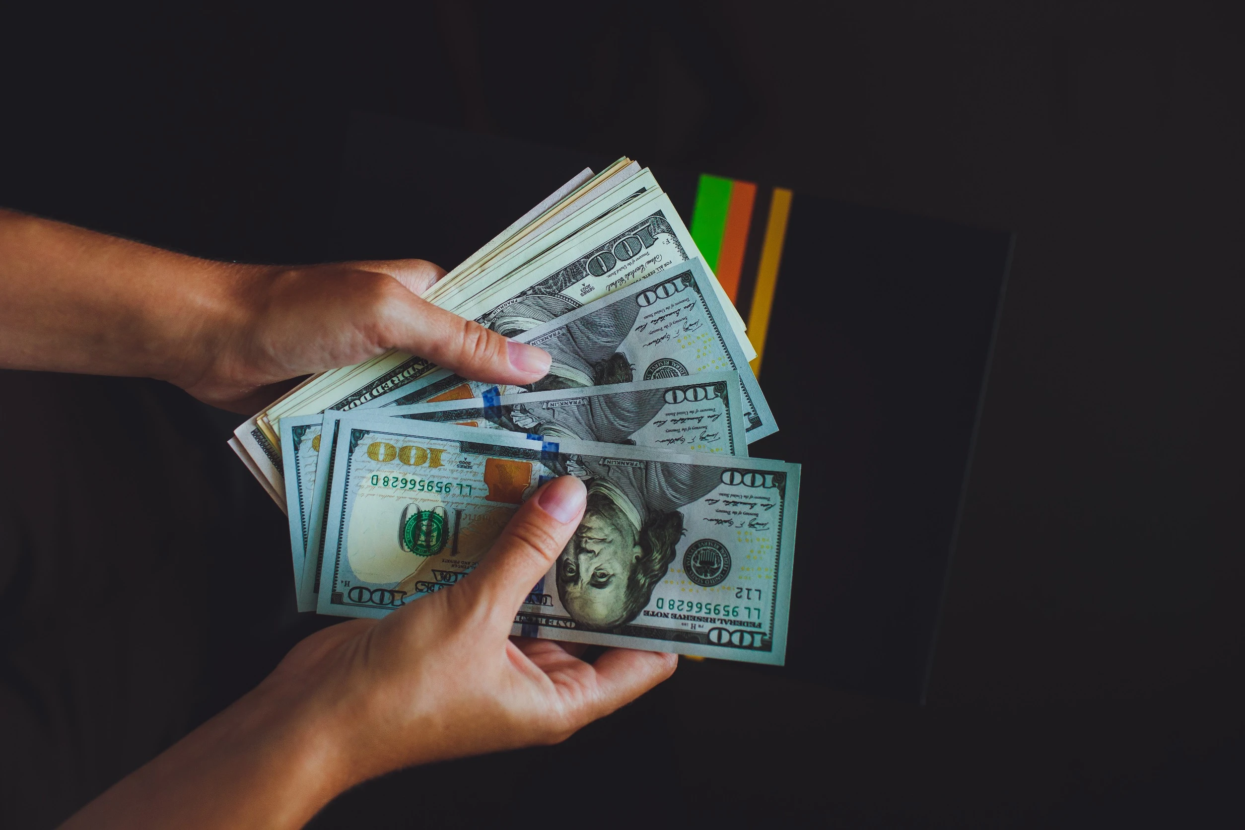 A pair of hands holding a stack of U.S. hundred-dollar bills, fanned out against a dark background with colorful stripes partially visible.
